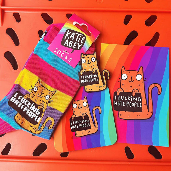 Contents of I F**king Hate People Sweary Cat Bundle on orange background. This includes an A6 print, coaster, magnet and stripy adult socks. All products are rainbow in colour and have an orange cat illustration holding a black sign with white writing on that reads 'I f**cking hate people'.