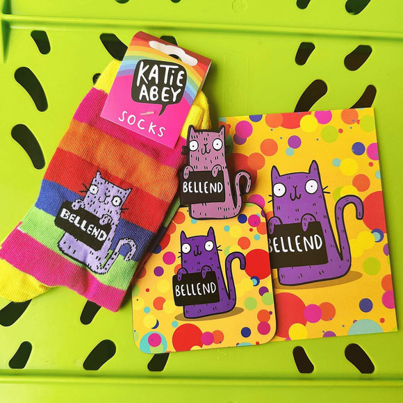 Contents of Bellend Sweary Cat Bundle on lime green background. Contents includes an a6 print, coaster, magnet, and rainbow stripe adult socks. All products are rainbow coloured with purple cat illustration holding a black sign with 'bellend' written in write letters.