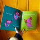 open book with one forrest green page with an illustration by Katie Abey of a pink axolotl in a shirt and tie using a fax machine with text 'fax-olotl' next to it. Underneath is an axolotl in a purple onesie eating crisps with text next to it 'snacks-olotl'. The lime green page next to it has an illustration of an axolotl playing the saxophone with text above 'groovy jazzy sax-olotl'