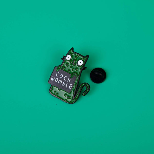 a soft enamel pin of a green smiley cat holding a black sign with white text on that reads cock womble. Designed by Katie Abey in the UK.