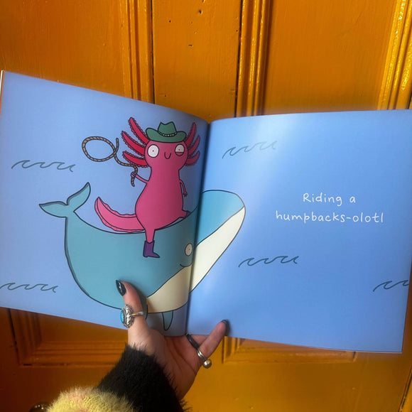 open book with blue pages and an illustration by Katie Abey on a pink smiling axolotl with a twitch in it's eye ,a cowboy hat on and is holding a lasso whilst riding a smiling whale across waves. Text reads 'riding a humpbacks-olotl'