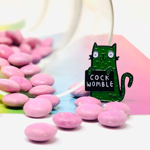 a soft enamel pin of a green smiley cat holding a black sign with white text on that reads cock womble. Designed by Katie Abey in the UK.