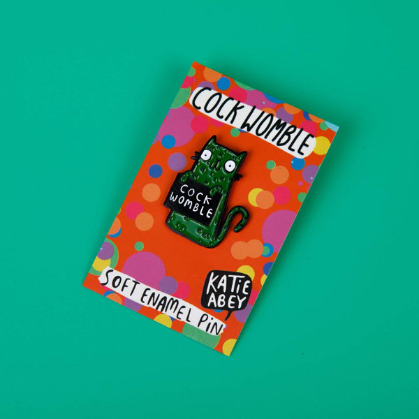 a soft enamel pin of a green smiley cat holding a black sign with white text on that reads cock womble. The pin is attached to its orange backing card with rainbow polka dots on. Designed by Katie Abey in the UK.