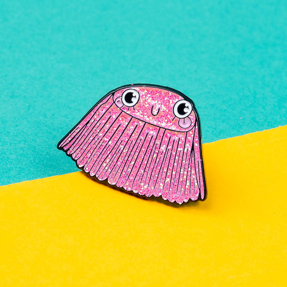 The Barnacle of Self Belief Enamel Pin Badge on a yellow and teal blue background. A pink glittering smiling barnacle pin with big eyes, eyelashes and pink cheeks all with a black outline. Hand drawn design by Katie Abey.