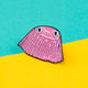 The Barnacle of Self Belief Enamel Pin Badge on a yellow and teal blue background. A pink glittering smiling barnacle pin with big eyes, eyelashes and pink cheeks all with a black outline. Hand drawn design by Katie Abey.