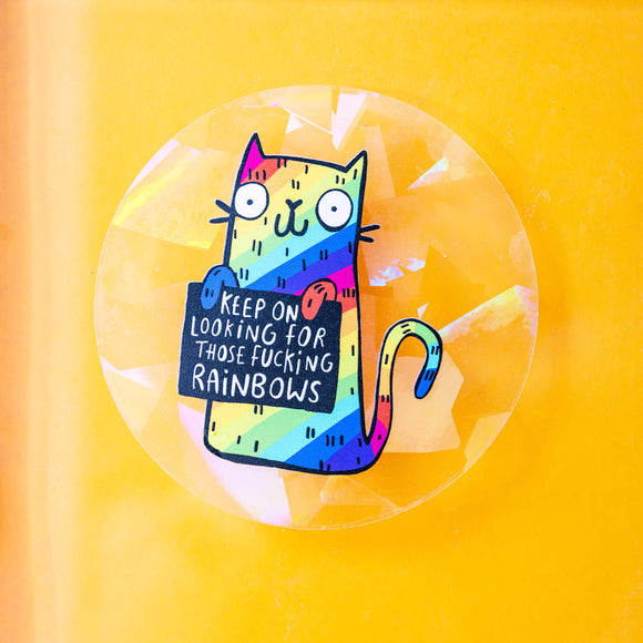 A see through rainbow making sun catching sticker that has a rainbow cat holding a sign saying keep on looking for those fucking rainbows