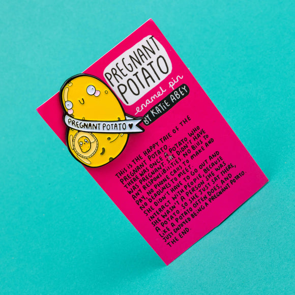 enamel pin badge shaped like a potato with a mini potato in its belly space and a banner around it that says pregnant potato. It is on a pink backing card telling the tale of the potato illustrated by Katie Abey