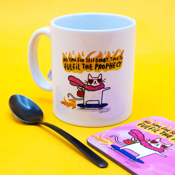 Side-angle view of a white Katie Abey mug with a fun and empowering cat illustration. The cat is zooming on a skateboard with fire behind it. Mug sits on a matching coaster with a spoon next to it on a yellow surface.