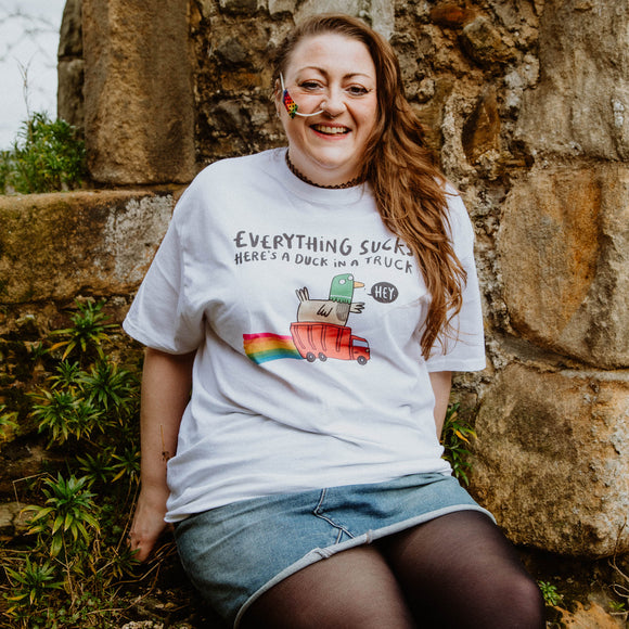 Everything Sucks Here's A Duck in a Truck black writing on a white cotton tshirt, featuring a waving green duck sitting in a red truck with rainbows illustration. Designed by Katie Abey in the UK.