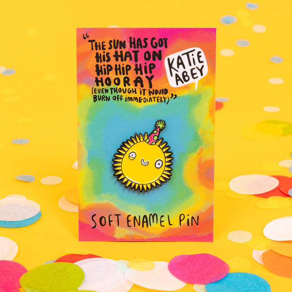 A soft enamel pin designed by Katie Abey with a sun smiling wearing a party hat it is on a backing card saying the sun has got his hat on hip hip hip hooray (even though it would definitely burn off immediately)