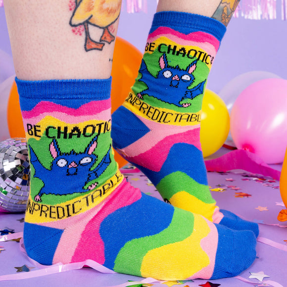 A pair of socks worn by a white model with tattoos. The socks have wavy multi coloured chunks and a drawing by Katie Abey of a bat with text saying be chaotic & Unpredictable