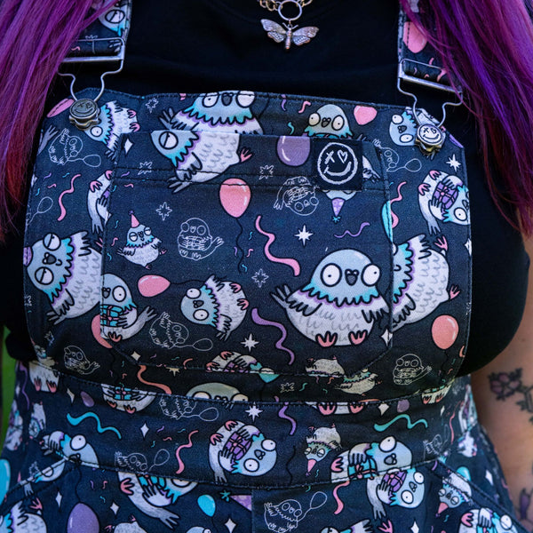 Close up of the Rebel Romance Ghost Party Flared Pinafore Dress showing the bib pocket, adjustable metal buckles and playful ghost print with balloons, stars and streamers. The dark cotton fabric features pastel pink, blue and lilac details for a fun alternative fashion look.
