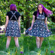 Front and back views of the Rebel Romance Ghost Party Flared Pinafore Dress worn outdoors. The ethical cotton pinafore features adjustable crossover straps, a flattering fitted waist, flared skirt and all over ghost party print with colourful balloons. Styled with a black T shirt for an alternative everyday outfit.