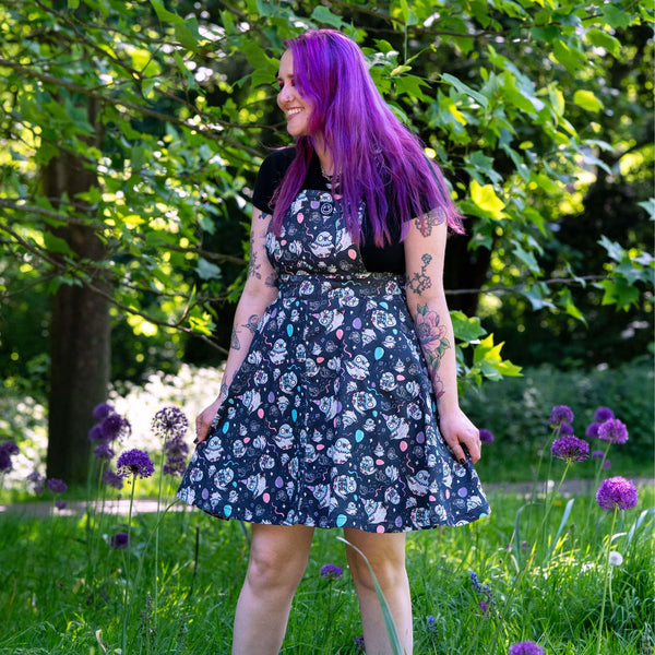 Model wearing the Rebel Romance Ghost Party Flared Pinafore Dress in a woodland garden. The dark cotton pinafore features adorable ghost characters, pastel balloons and confetti details, with adjustable straps, a bib pocket and flared skirt. A fun size inclusive alternative fashion dress perfect for everyday wear or spooky season.