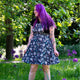 Model wearing the Rebel Romance Ghost Party Flared Pinafore Dress in a woodland garden. The dark cotton pinafore features adorable ghost characters, pastel balloons and confetti details, with adjustable straps, a bib pocket and flared skirt. A fun size inclusive alternative fashion dress perfect for everyday wear or spooky season.