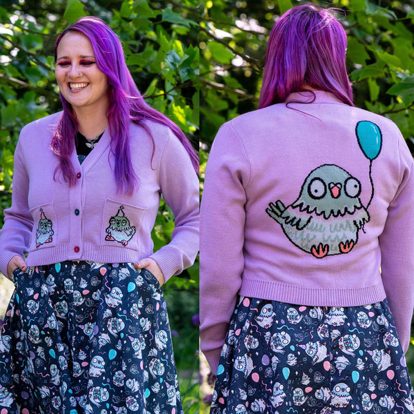 Model wearing the Rebel Romance x Katie Abey Pigeon Party Cardigan in lilac with embroidered party pigeons on the front pockets. Styled over the matching Pigeon Party Flared Pinafore Dress, this soft vegan friendly cardigan features colourful buttons, long sleeves and a cropped fit. Perfect for colourful alternative fashion lovers.
