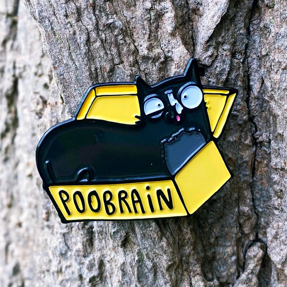 The Poobrain Cat Enamel Pin on a tree trunk. The enamel pin is of an open yellow box with a black cat inside with a silly expression. There is black text on the box that reads POOBRAIN. Designed by Katie Abey in the UK