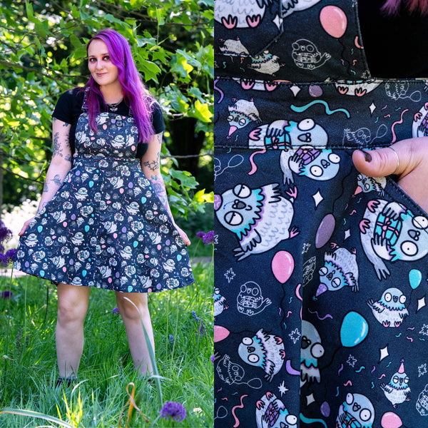 Model with bright purple hair wearing the Rebel Romance Ghost Party Flared Pinafore Dress in a garden setting. This ethical alternative dress features a cute ghost and pastel balloon print, adjustable straps, bib pocket and a twirly flared skirt. Close up image highlights the colourful spooky print and practical side pocket.