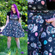 Model with bright purple hair wearing the Rebel Romance Ghost Party Flared Pinafore Dress in a garden setting. This ethical alternative dress features a cute ghost and pastel balloon print, adjustable straps, bib pocket and a twirly flared skirt. Close up image highlights the colourful spooky print and practical side pocket.