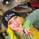 Katie Abey wearing the black Embracing The Chaos Cat Beanie Hat by Katie Abey standing in front of a dinosaur sculpture. Katie has bright yellow hair and colourful accessories. The beanie shows the embroidered chaotic cat and text Embracing The Chaos. Cosy ethical winter hat for quirky style lovers.