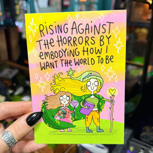 A6 illustrated postcard by Katie Abey featuring two whimsical characters standing together under a pink and yellow sky with sparkles. One holds a glowing heart wand and book, the other clutches a small object, both wearing green cloaks. Handwritten text reads “Rising against the horrors by embodying how I want the world to be”. Bright, magical and empowering artwork printed on silk card.