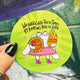 Bright green circular vinyl sticker illustrated by Katie Abey featuring a cute white character holding a sunflower labelled magic and whimsy and a flaming heart labelled political outrage. Handwritten text reads we navigate these times by keeping hold of both. Bold, playful illustration with sparkles, pink skirt and boots, celebrating balance, feelings and being unapologetically weird.