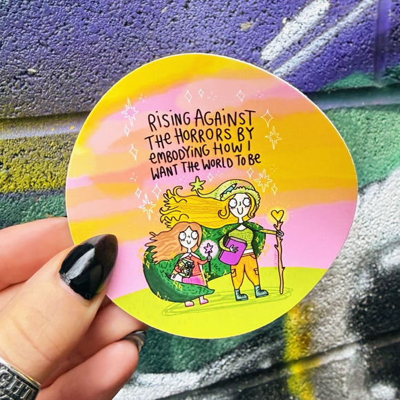 Round vinyl sticker by Katie Abey featuring a colourful illustrated adult and child holding magical wands, standing together against a pastel pink, yellow and green sky. The sticker includes the handwritten quote “Rising against the horrors by embodying how I want the world to be”. A joyful, empowering design encouraging kindness, creativity and being unapologetically weird.