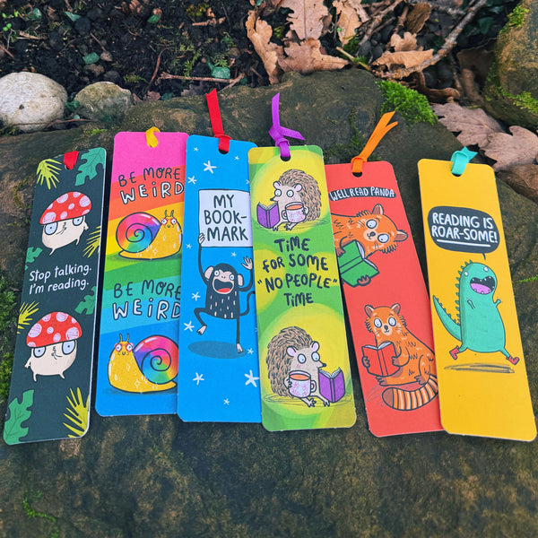 Katie Abey Stop Talking, I’m Reading mushroom bookmark displayed with other colourful illustrated bookmarks outdoors on a mossy rock. Features a quirky red toadstool character on a dark green background with white text and ribbon tie. Sturdy laser printed card with silk finish. Cute gift for bookworms and stationery lovers.

