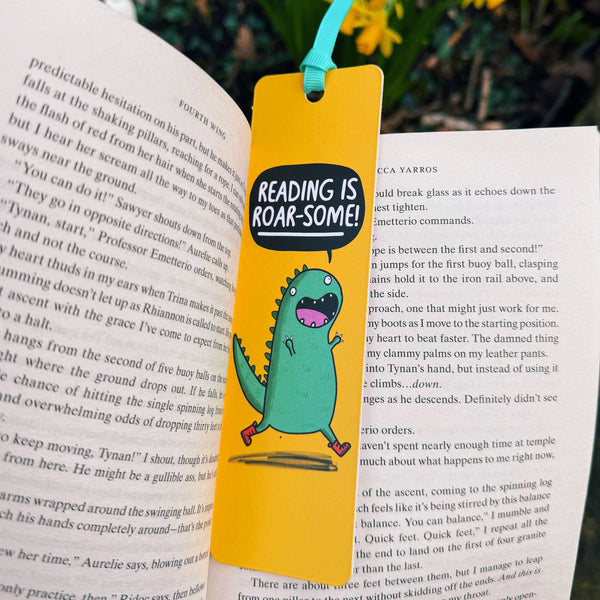 Reading Is Roar-Some Dinosaur Bookmark by Katie Abey placed inside an open book outdoors. Bright yellow bookmark with cute green dinosaur illustration and speech bubble text. Ribbon tie at the top. Laser printed on sturdy card with premium silk finish. Fun and quirky gift for book lovers who adore reading and embracing their weird side.