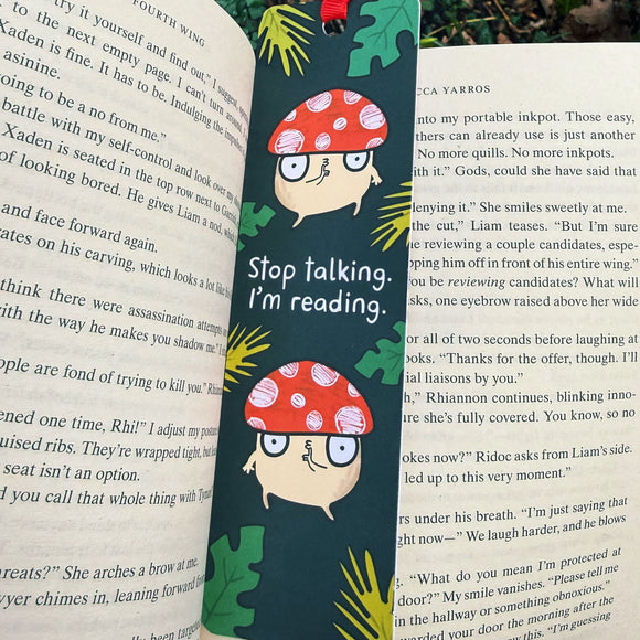 Stop Talking, I’m Reading mushroom bookmark by Katie Abey placed inside an open book. Dark green background with quirky red toadstool characters and bold white slogan text. Laser printed on sturdy card with a premium silk finish and red ribbon tie. Cute illustrated bookmark for book lovers, readers and fans of fun stationery.
