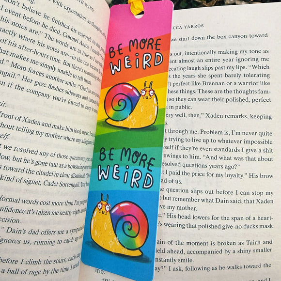 Be More Weird Snail Bookmark by Katie Abey inside an open book, featuring a rainbow background and cute illustrated snail with slogan. Laser printed on sturdy card with silk finish and ribbon tie, colourful quirky bookmark for book lovers and stationery fans.
