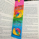 Be More Weird Snail Bookmark by Katie Abey inside an open book, featuring a rainbow background and cute illustrated snail with slogan. Laser printed on sturdy card with silk finish and ribbon tie, colourful quirky bookmark for book lovers and stationery fans.
