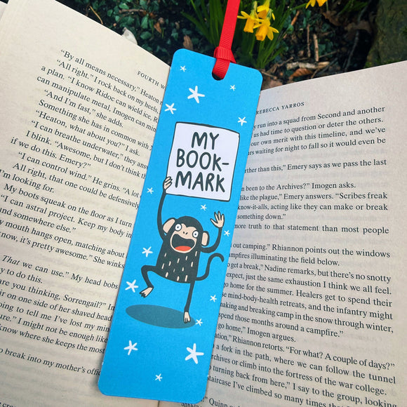 Blue “My Bookmark” Monkey Bookmark by Katie Abey placed inside an open book outdoors. The illustrated monkey holds a sign reading “My Book-mark” against a starry blue background with a red ribbon tie. Laser printed on sturdy card with a premium silk finish. A colourful, quirky bookmark from small UK illustrator Katie Abey, perfect for book lovers who like to be more weird.
