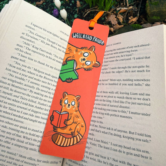 Well Read Panda red panda bookmark by Katie Abey placed inside an open book outdoors. Bright red bookmark featuring cute illustrated red pandas reading green books with ribbon tie top. Laser printed on sturdy card with premium silk finish. Quirky book lover gift from UK illustrator encouraging you to be more weird.
