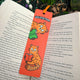 Well Read Panda red panda bookmark by Katie Abey placed inside an open book outdoors. Bright red bookmark featuring cute illustrated red pandas reading green books with ribbon tie top. Laser printed on sturdy card with premium silk finish. Quirky book lover gift from UK illustrator encouraging you to be more weird.
