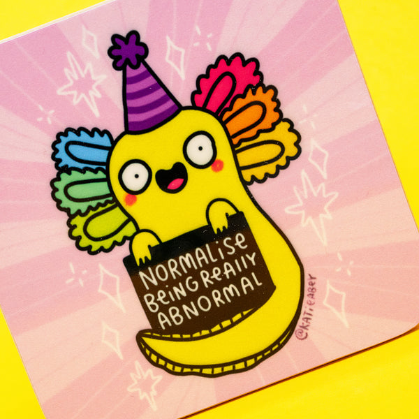 A close up of the Normalise Being Really Abnormal Axolotl Coaster by Katie Abey. The happy yellow axolotl with rainbow gills and a striped party hat holds a playful sign on a pink starburst background. The coaster has rounded edges and is photographed against a yellow backdrop, ideal for anyone who loves fun, weird, uplifting home decor.