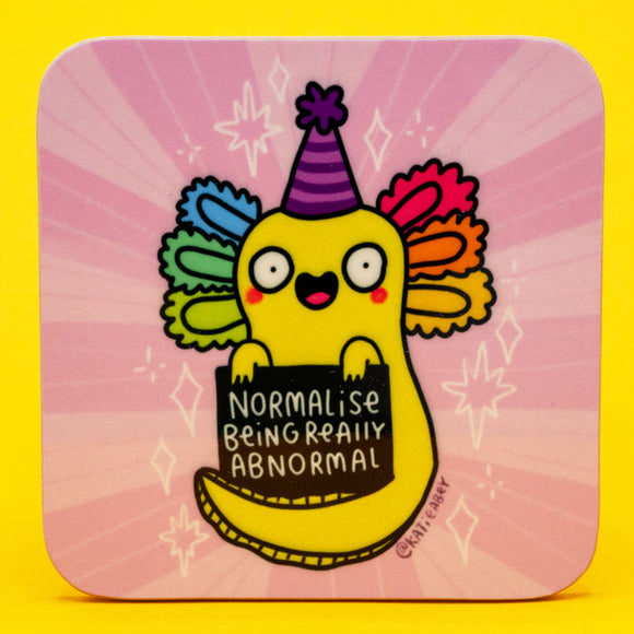 A bright pink square coaster featuring a cheerful yellow axolotl illustrated by Katie Abey. The axolotl wears a purple party hat and rainbow gills while holding a sign that says Normalise Being Really Abnormal. The coaster has rounded edges and sits on a yellow background. Cute, quirky, and perfect for fans of weird and colourful homeware.