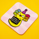 The Normalise Being Really Abnormal Axolotl Coaster by Katie Abey shown at an angle on a yellow background. The bright yellow axolotl with rainbow gills, a purple party hat, and a positive message sits on a pink illustrated coaster with rounded edges. A colourful, quirky design for fans of weird and wonderful accessories.