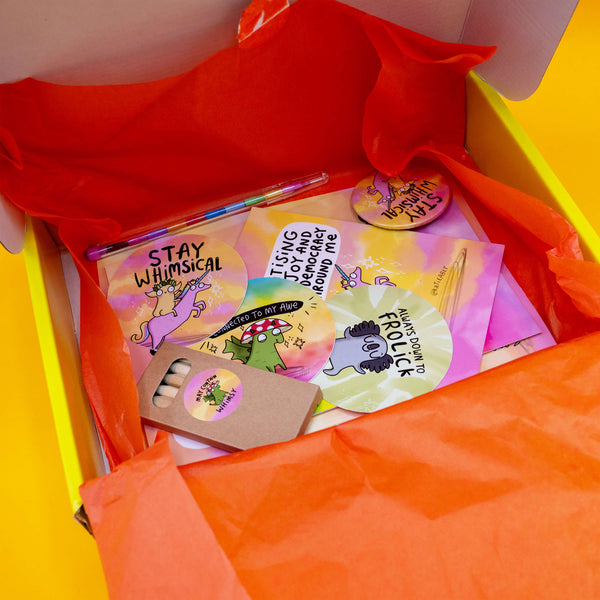 Inside view of the open prioritising whimsy themed Box of Brilliance subscription box. The yellow box lined with orange tissue paper and includes vinyl stickers, postcards, a badge, a mini colouring pencil set, and a rainbow pen. Features playful pastel designs with quirky characters and empowering quotes. A vibrant, joyful box filled with magical positivity.