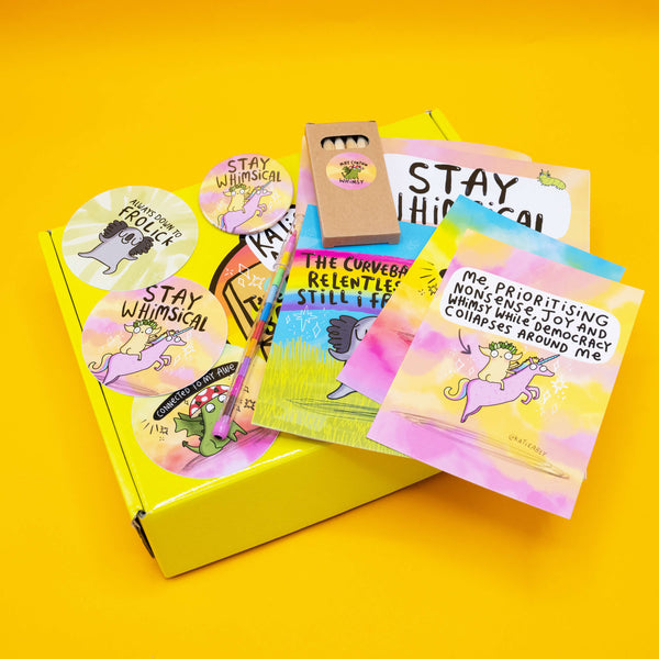 Flat lay of Katie Abey’s Prioritising Whimsy themed Box of Brilliance subscription box on a bright yellow background. The open yellow box contains a colouring book, postcards, vinyl stickers, a badge, a mini pencil set, and a rainbow pen. Visible designs include joyful characters like a unicorn, dragon, and koala with uplifting and humorous affirmations. A colourful, feel-good bundle of whimsy and magic.