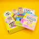 Flat lay of Katie Abey’s Prioritising Whimsy themed Box of Brilliance subscription box on a bright yellow background. The open yellow box contains a colouring book, postcards, vinyl stickers, a badge, a mini pencil set, and a rainbow pen. Visible designs include joyful characters like a unicorn, dragon, and koala with uplifting and humorous affirmations. A colourful, feel-good bundle of whimsy and magic.