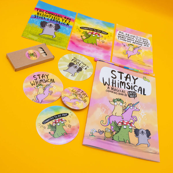 Flat lay of Katie Abey’s colourful “prioritising whimsy” product collection on a bright yellow background. Includes a magical colouring book, large and small vinyl stickers, a button badge, a mini pencil set, and postcard prints with whimsical characters and uplifting quotes. A playful mix of pastel tones, quirky creatures, and joyful affirmations from the UK illustrator encouraging positivity and weirdness.