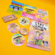 Flat lay of Katie Abey’s colourful “prioritising whimsy” product collection on a bright yellow background. Includes a magical colouring book, large and small vinyl stickers, a button badge, a mini pencil set, and postcard prints with whimsical characters and uplifting quotes. A playful mix of pastel tones, quirky creatures, and joyful affirmations from the UK illustrator encouraging positivity and weirdness.