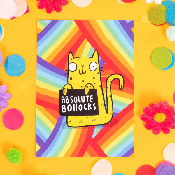 The Sweary Cat Absolute B*llocks A6 Postcard in front of a yellow backdrop with colourful flower and confetti decorations. The rainbow striped postcard has an illustration of a yellow smiling cat on the front holding a black sign with white text that reads absolute bollocks. Designed by Katie Abey in the UK.