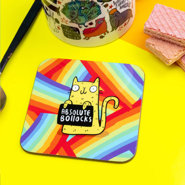 a coaster of a yellow cat on a rainbow striped background smiling holding a sign saying 'ABSOLUTE BOLLOCKS'