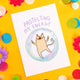 A white postcard with an illustration of a cat looking zen in a bubble with the words 'protecting my energy' above it. The postcard is on a yellow background with colourful confetti, fake daisies and pom poms 