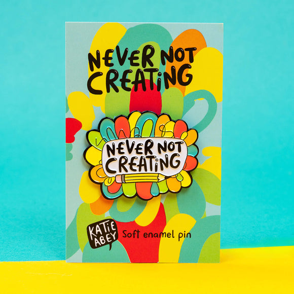 Katie Abey soft enamel pin featuring the motivational phrase 'Never Not Creating' surrounded by a colourful design. The pin is displayed on a bright backing card with the same phrase at the top and the artist's name, Katie Abey, at the bottom. Perfect for inspiring artists and creatives.