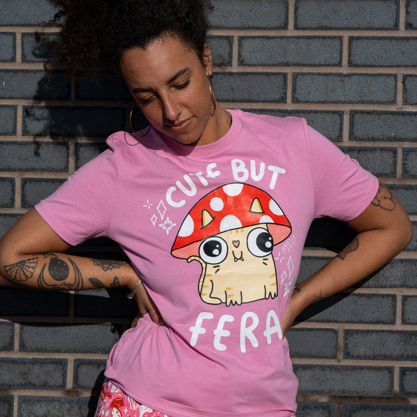 Front view of the Cute But Feral Cat Adults Tee in bubble pink by Katie Abey. The pink t shirt features a cute illustrated cat wearing a red spotted mushroom hat with the slogan Cute But Feral. Model stands against a blue brick wall wearing pink mushroom print trousers. Fun illustrated clothing by a small UK business.