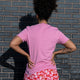 Back view of a model wearing the Cute But Feral Cat Adults Tee in bubble pink. The soft pink t shirt has a relaxed unisex fit and is styled with pink mushroom print bottoms. Photograph taken outdoors against a blue brick wall. Designed by Katie Abey, an illustrator known for quirky, cute and weird artwork.
