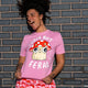 Model wearing the Cute But Feral Cat Adults Tee in bubble pink, smiling and posing outdoors against a blue brick wall. The pink t shirt shows a playful illustrated feral cat with mushroom hat and bold white text. Designed by Katie Abey, a small business illustrator encouraging self expression and embracing being weird.
